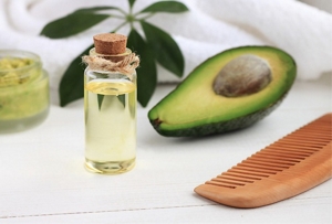 Bottle of avocado oil sitting on a white table alongside a comb and an avocado